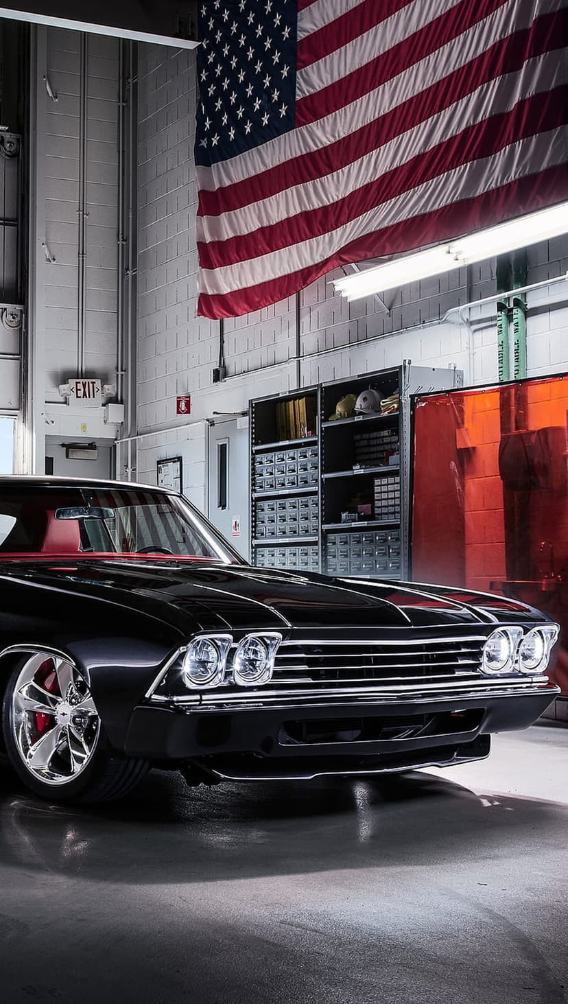 Classic black muscle car with a red interior parked in a garage under a large American flag with chrome wheels.