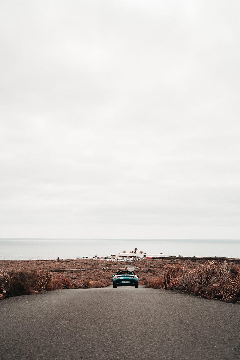 Blue convertible car driving down a remote coastal road toward a vast ocean horizon under an overcast gray sky.