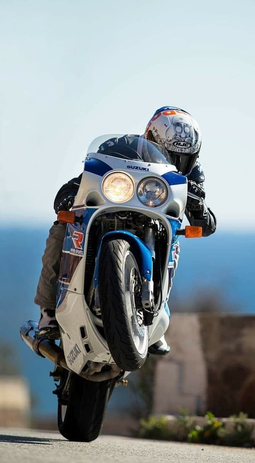 White and blue Suzuki motorcycle mid-wheelie on a scenic coastal road with the rider in a skull helmet.