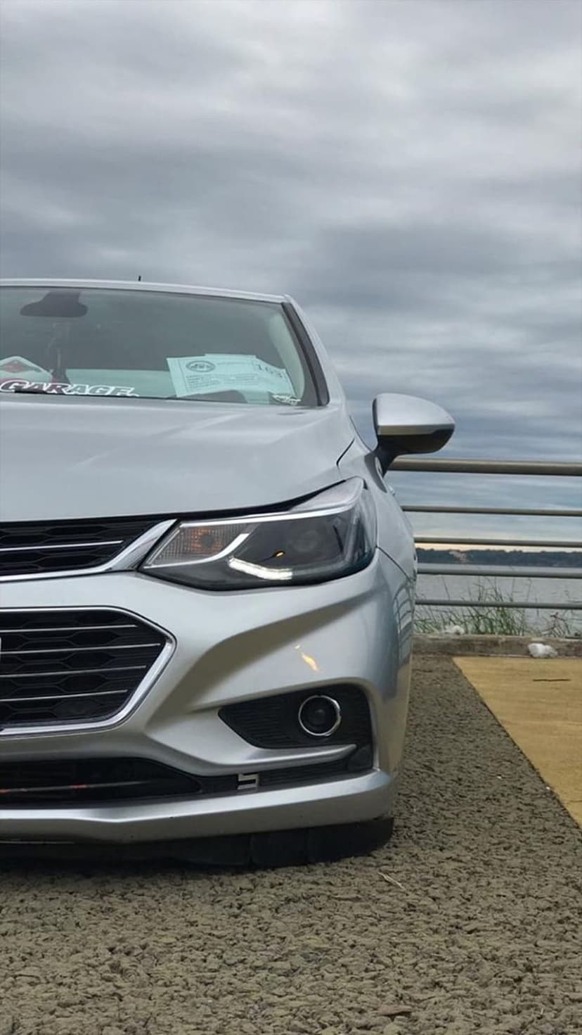 Silver Chevrolet Cruze front grille and headlight parked near a railing with a cloudy sky and water background.