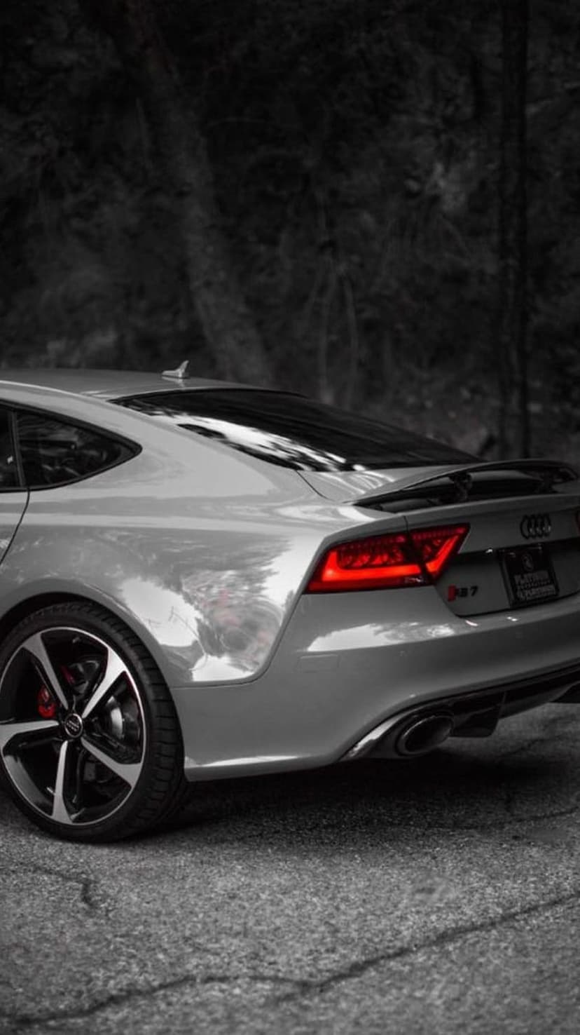 Silver Audi RS7 parked on an asphalt road with glowing red taillights and dark trees in the blurred background.