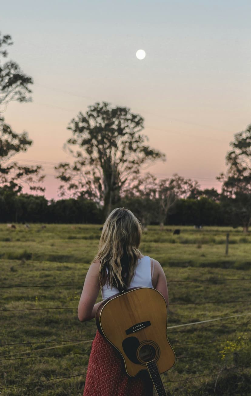 Woman holding a guitar faces a pastel sunset and full moon while standing in a grassy meadow during twilight.