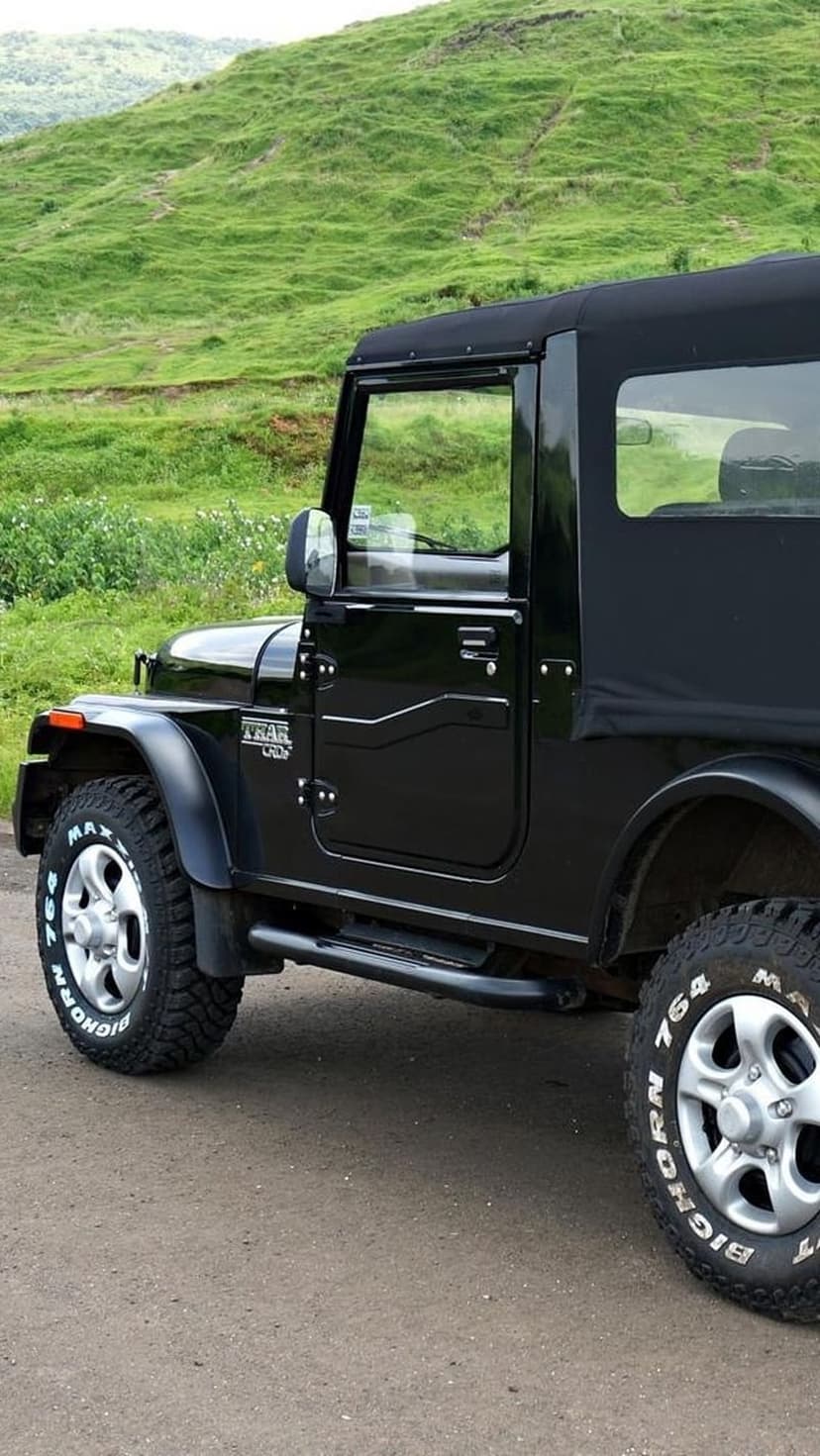 Black Mahindra Thar SUV parked on a dirt path with chunky off-road tires and a backdrop of lush green hills.
