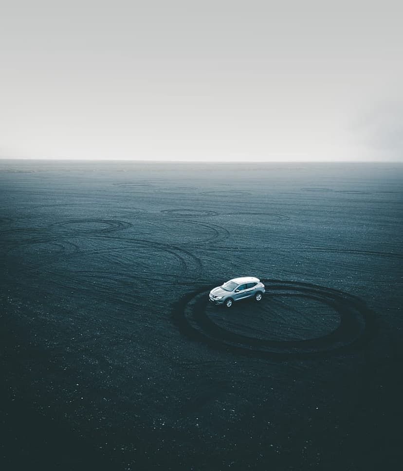 Silver SUV carves circular tire tracks on a black sand beach viewed from a high-angle aerial drone perspective.