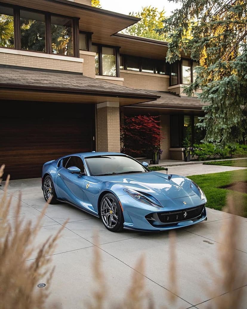 Blue Ferrari supercar parked on a driveway before a multi-story glass and brick modern luxury residence.