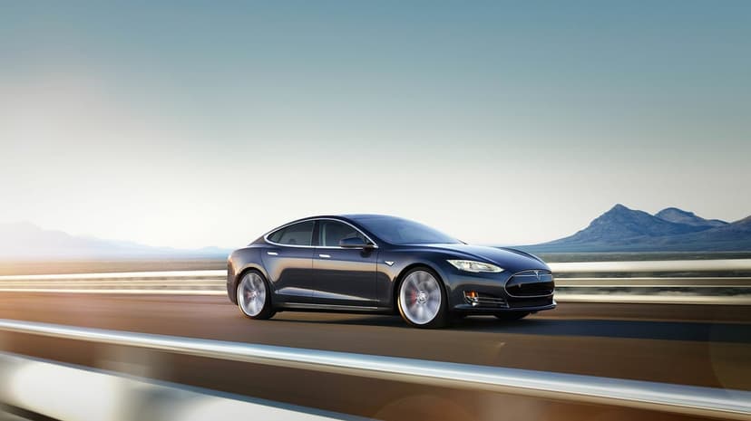 Dark grey Tesla Model S driving fast on a desert highway with mountain peaks and a clear sky background.