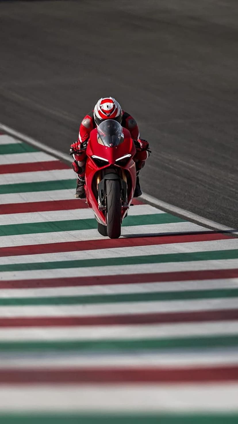 Red Ducati motorcycle rider leans into a racetrack turn near a tricolore striped kerb with blurred asphalt.