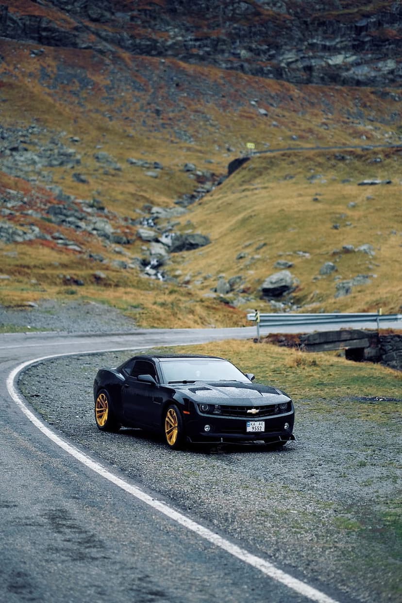 Black Chevrolet Camaro with gold rims parked on a winding mountain road among rocky slopes and autumn vegetation.