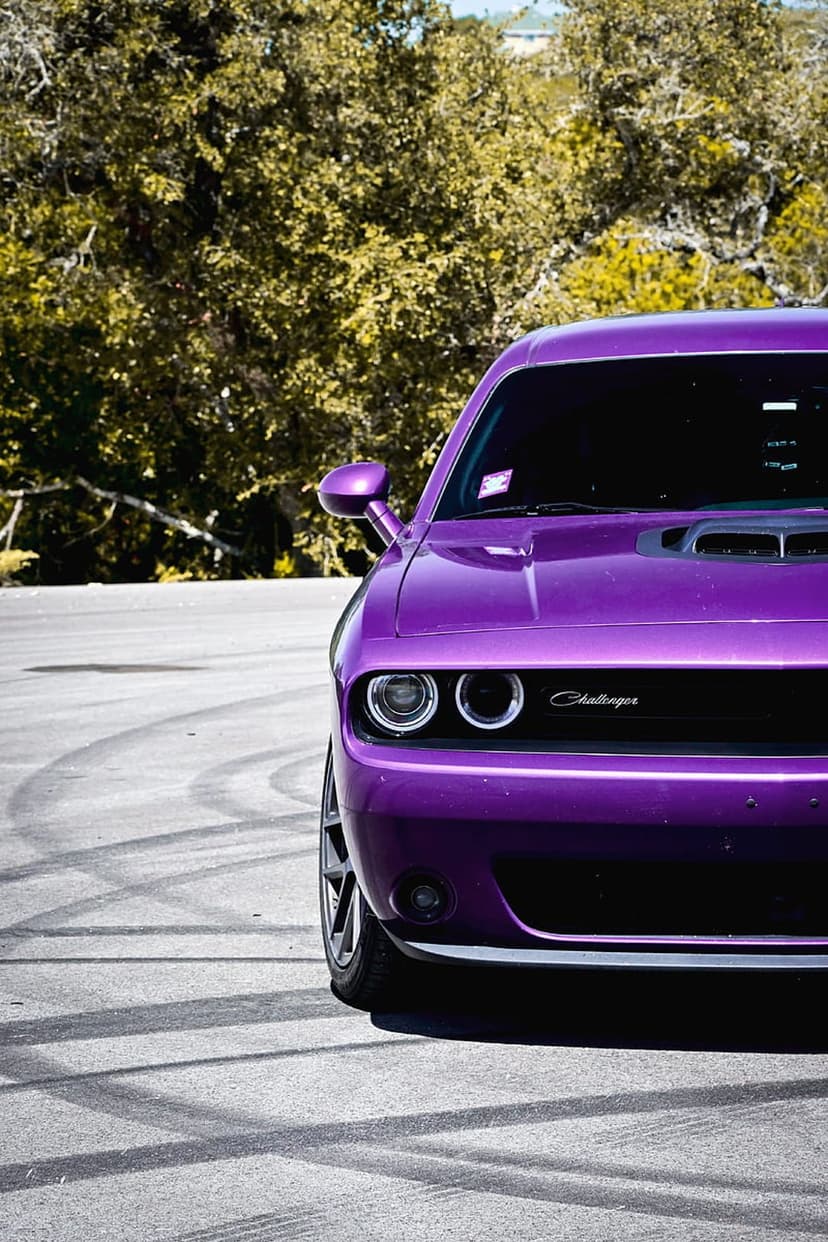 Purple Dodge Challenger showing its front grille and headlights on asphalt with skid marks and green trees.