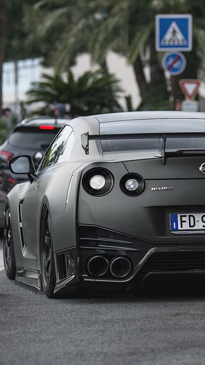Nissan GT-R Nismo in matte gray shows circular taillights and a carbon fiber spoiler on a paved road with trees.