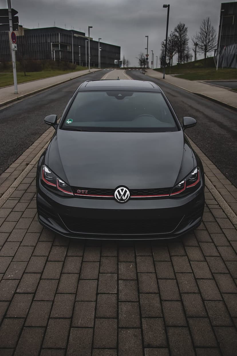 Dark grey Volkswagen Golf GTI parked on a brick road with a red grille stripe under an overcast urban sky.