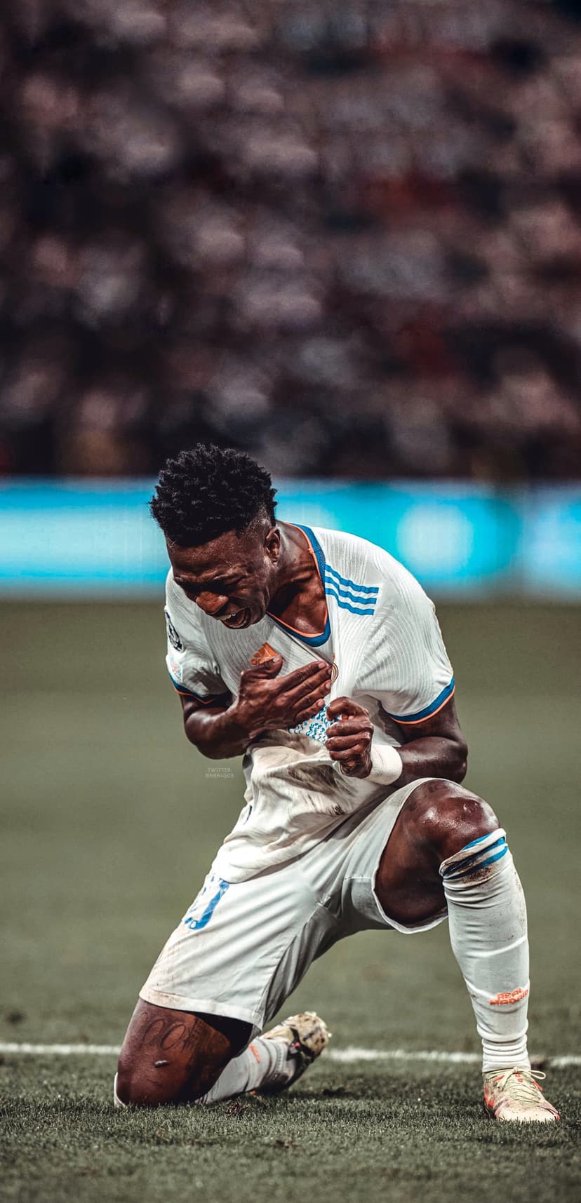 Vinicius Junior kneels on a green football pitch while clutching his white Real Madrid jersey in celebration.
