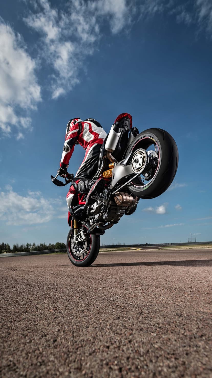 Racer in full gear lifts a motorcycle into a vertical wheelie against a bright blue sky with white clouds.