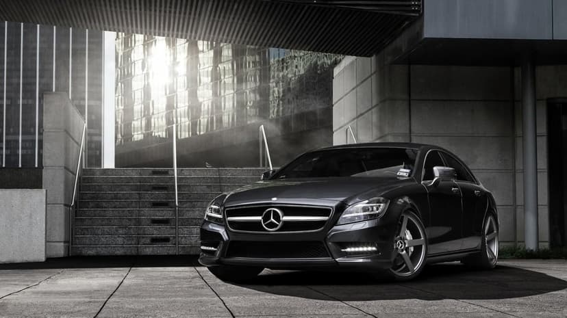 Gunmetal grey Mercedes-Benz CLS parked by concrete stairs with bright sunlight reflecting off a glass building.