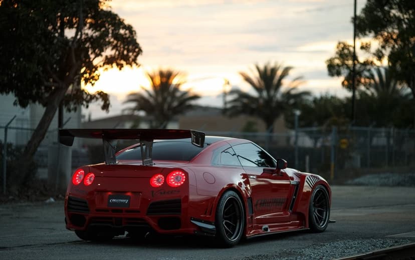 Red Nissan GT-R with widebody kit and massive rear wing parked on asphalt at sunset with glowing taillights.