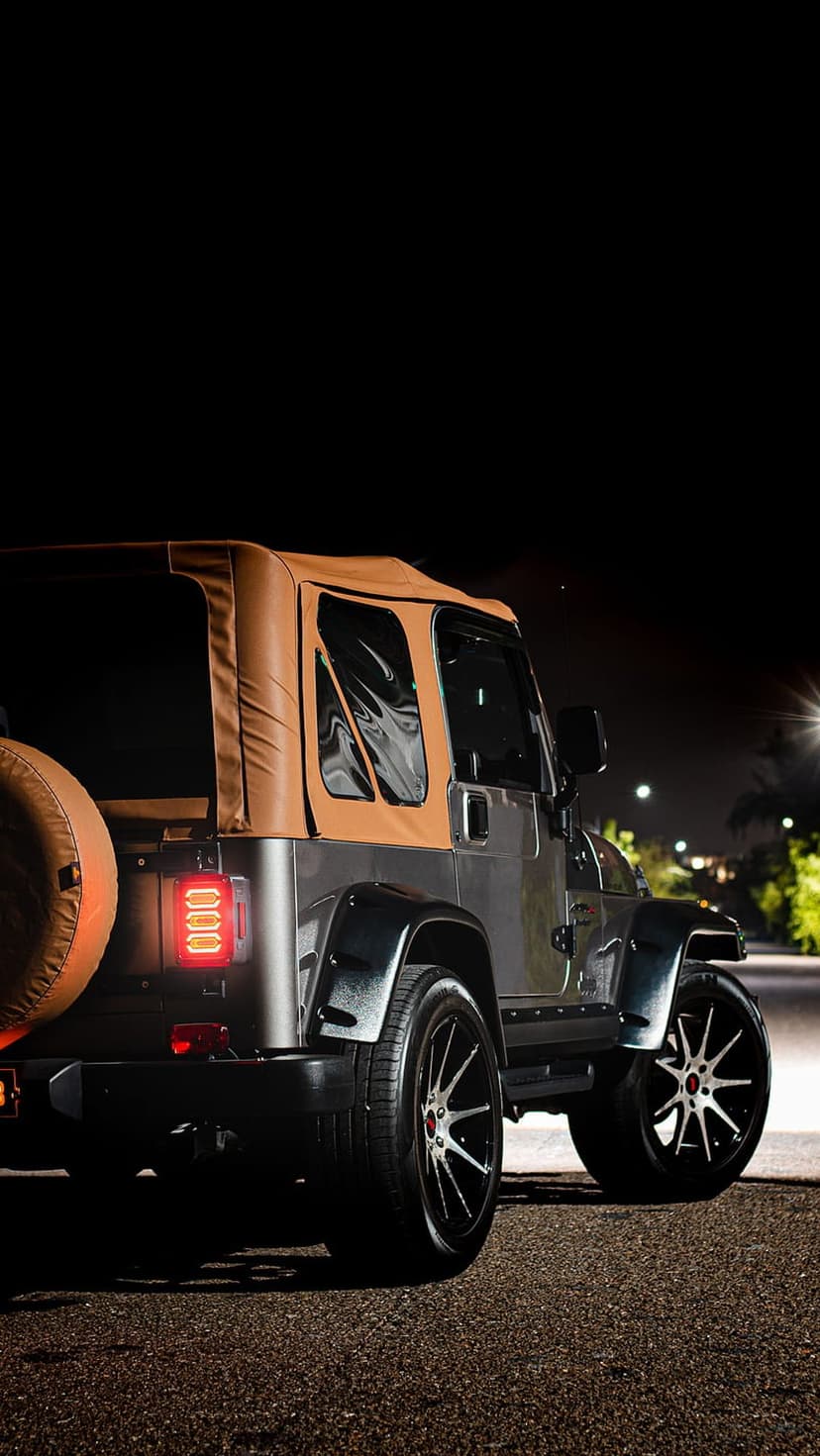 Jeep Wrangler SUV with a tan soft top and red-accented wheels parked on asphalt during a dark urban night.