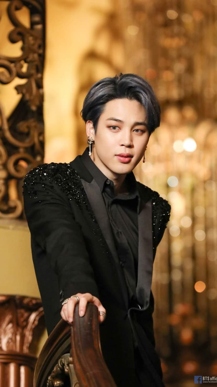 Jimin of BTS wears a black suit with sparkling shoulders while leaning on a wooden rail with silver drop earrings.