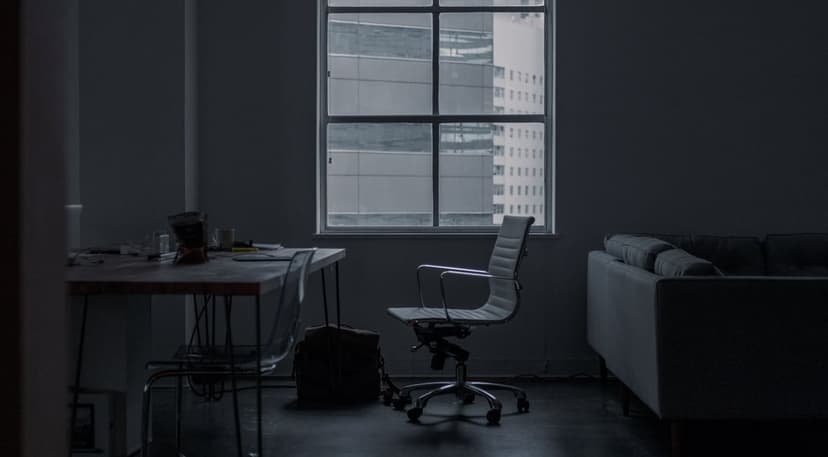 Sleek desk with a laptop and ergonomic chair in a dim room against a window with a blurred urban cityscape view.