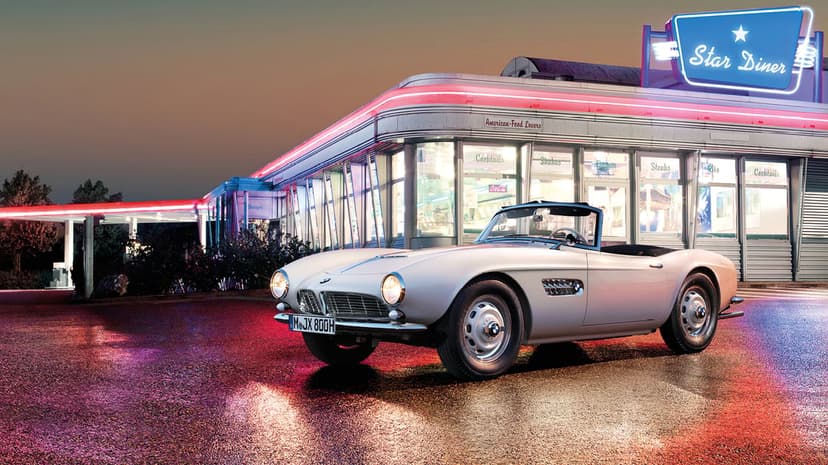 Silver vintage BMW 507 roadster parked in front of a Star Diner with red and blue neon lighting reflections.