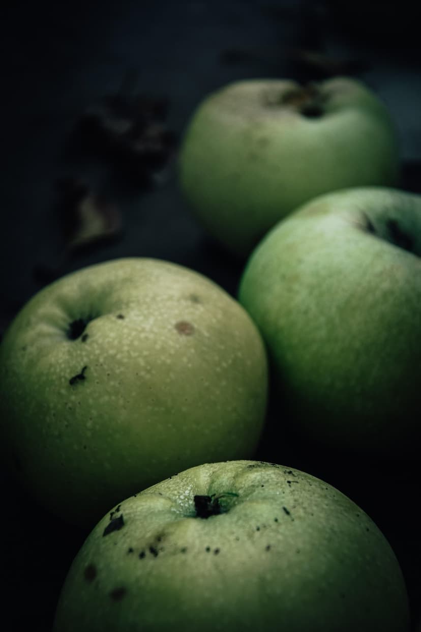 Ripe green apples with visible textures and blemishes sit on a dark surface under atmospheric lighting.
