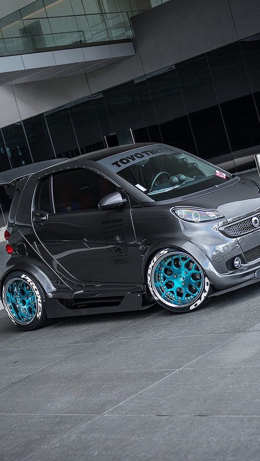 Dark grey Smart car with teal multi-spoke wheels and a rear spoiler parked near a modern glass building.