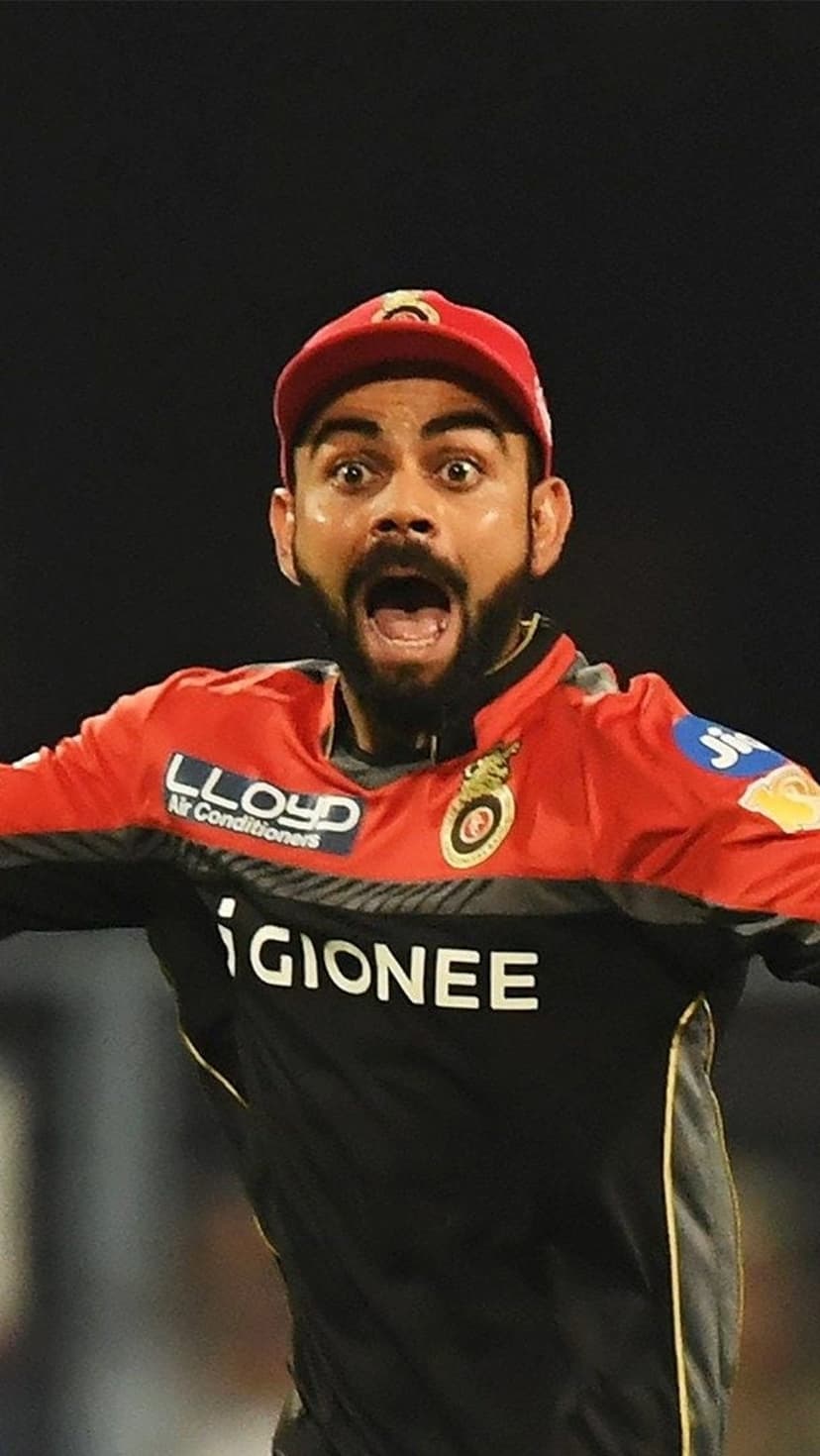 Virat Kohli wears a red RCB cap and jersey with a surprised open-mouthed expression during an IPL cricket match.