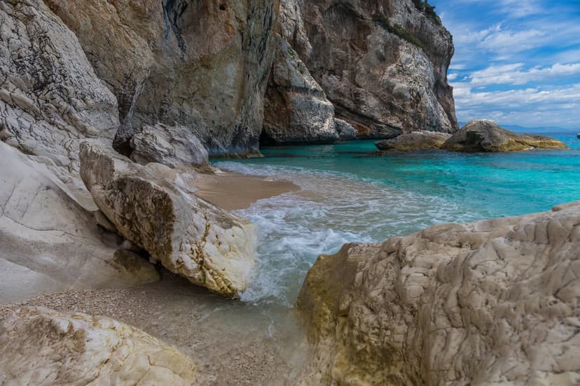 Limestone cliffs tower over a secluded turquoise cove with clear water washing onto a pale sandy beach at midday.