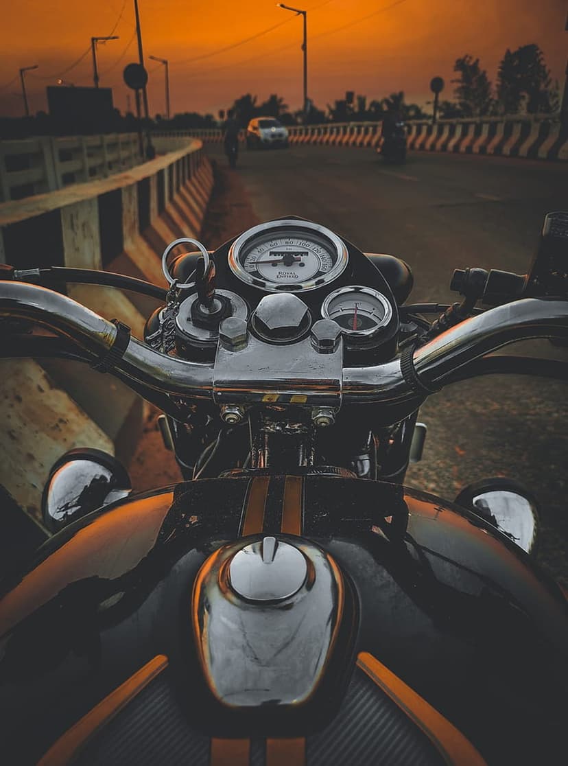 Motorcycle speedometer and handlebars reflect a glowing orange sunset sky during a highway ride at twilight.