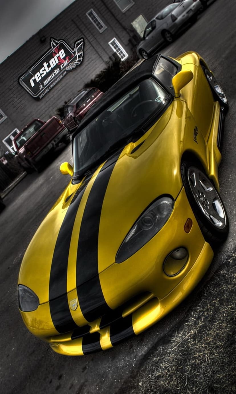 Yellow Dodge Viper convertible with black racing stripes parked in front of a muscle car restoration building.