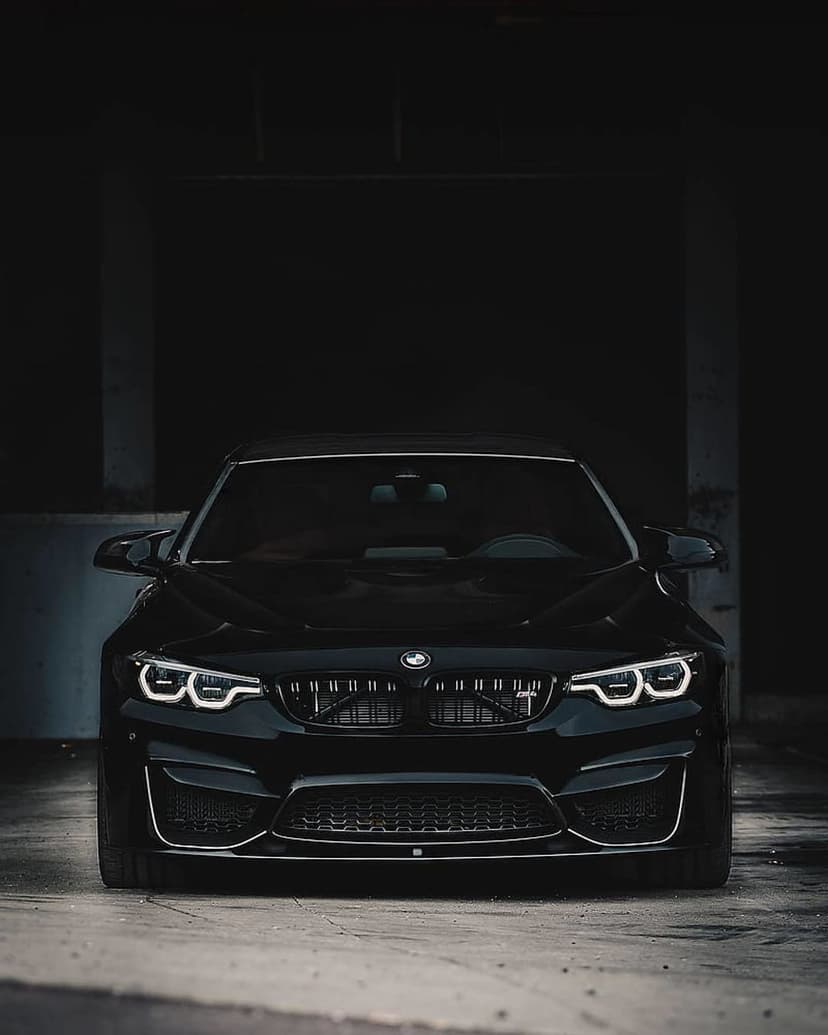 Black BMW M4 Coupe with illuminated LED headlights and a large kidney grille parked in a dark garage.