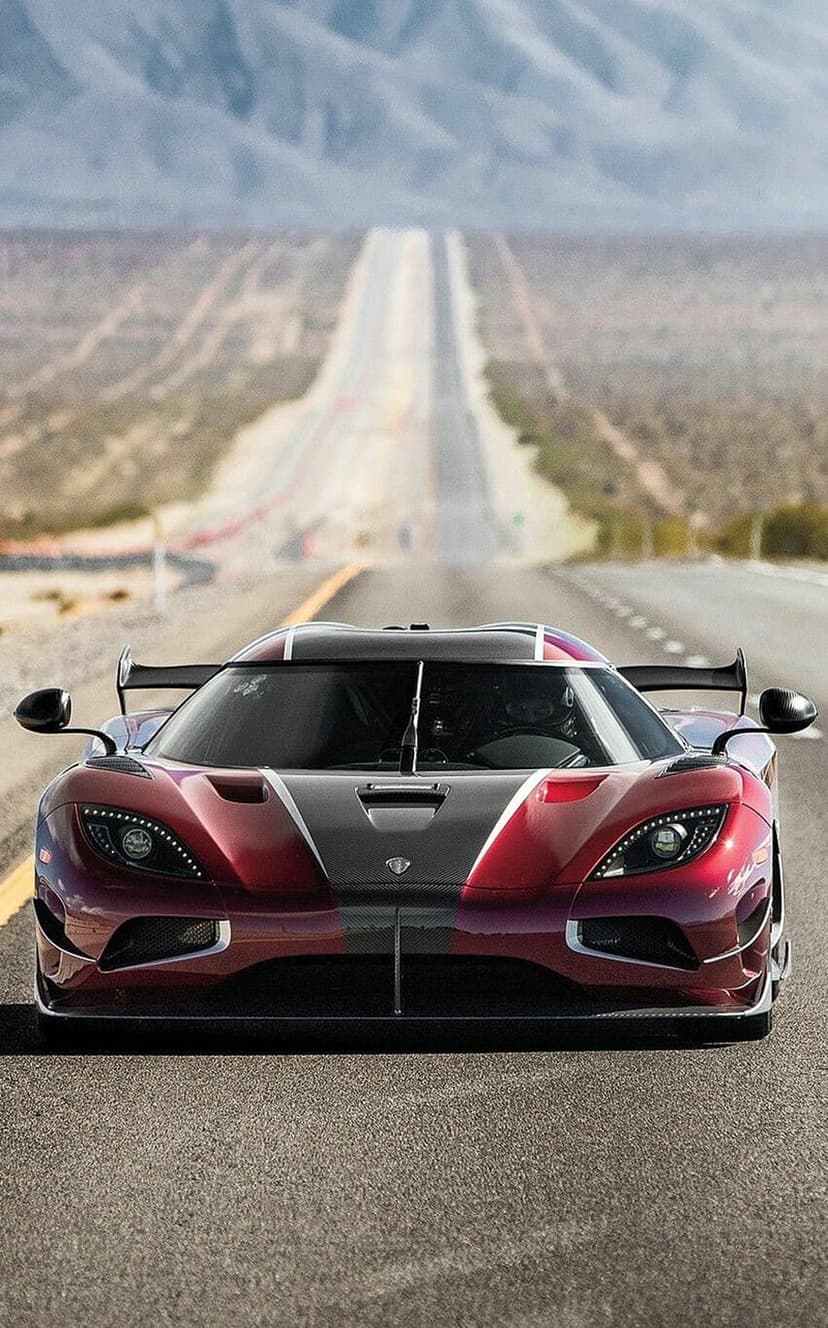 Red Koenigsegg hypercar speeding on a straight desert road with rugged mountains under a bright and clear sky.