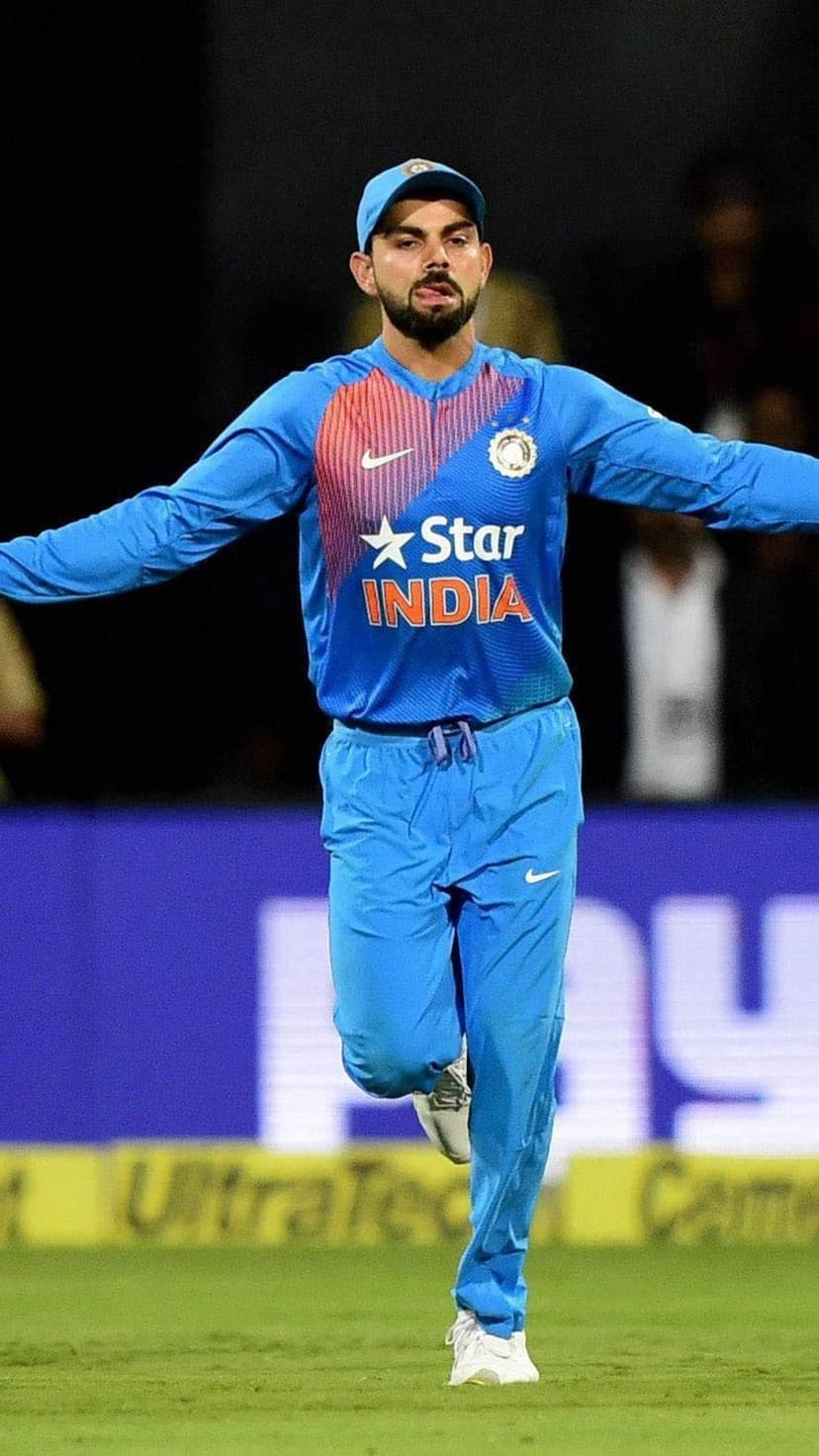 Virat Kohli sprints across a green cricket pitch wearing a blue India jersey with his arms outstretched in celebration.