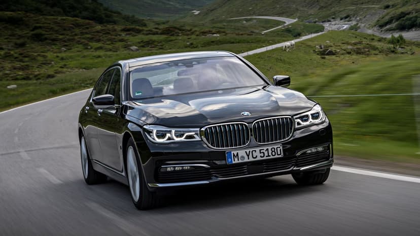 Black BMW 7 Series sedan driving on a winding mountain road through green hills under an overcast sky.