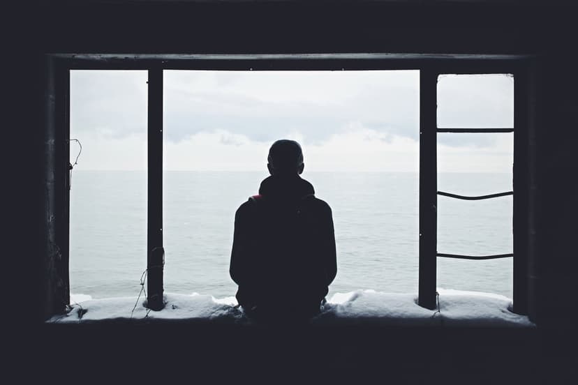 Solitary silhouette standing by a snow-covered window frame overlooking a vast, dark ocean under a cloudy sky.