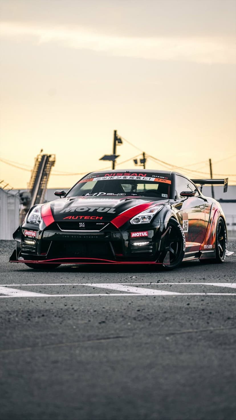 Black Nissan GT-R with red stripes parked on asphalt under an orange sunset sky near industrial buildings.
