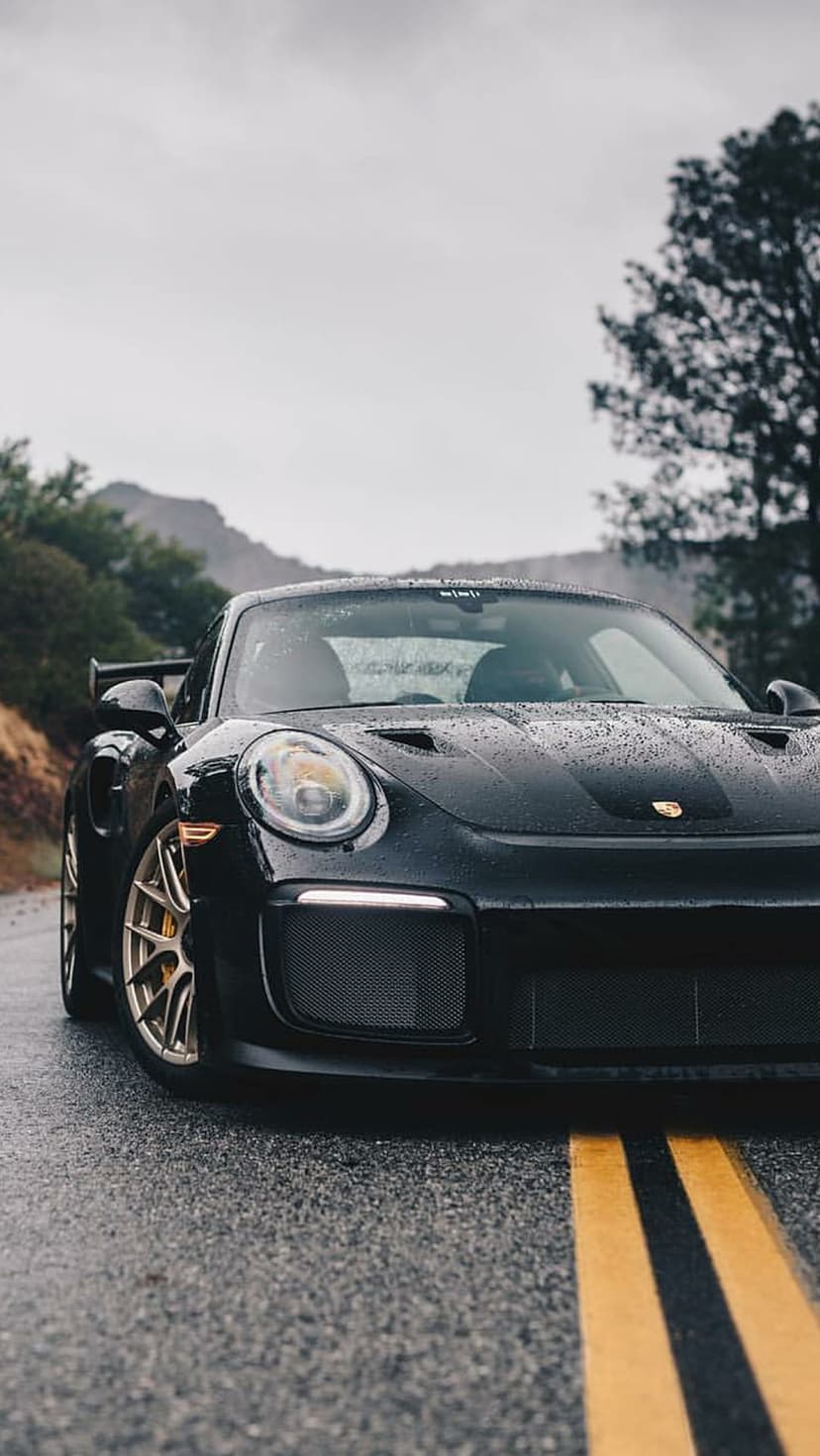 Black Porsche 911 GT2 RS with raindrops on a wet asphalt road against a misty mountain forest backdrop.
