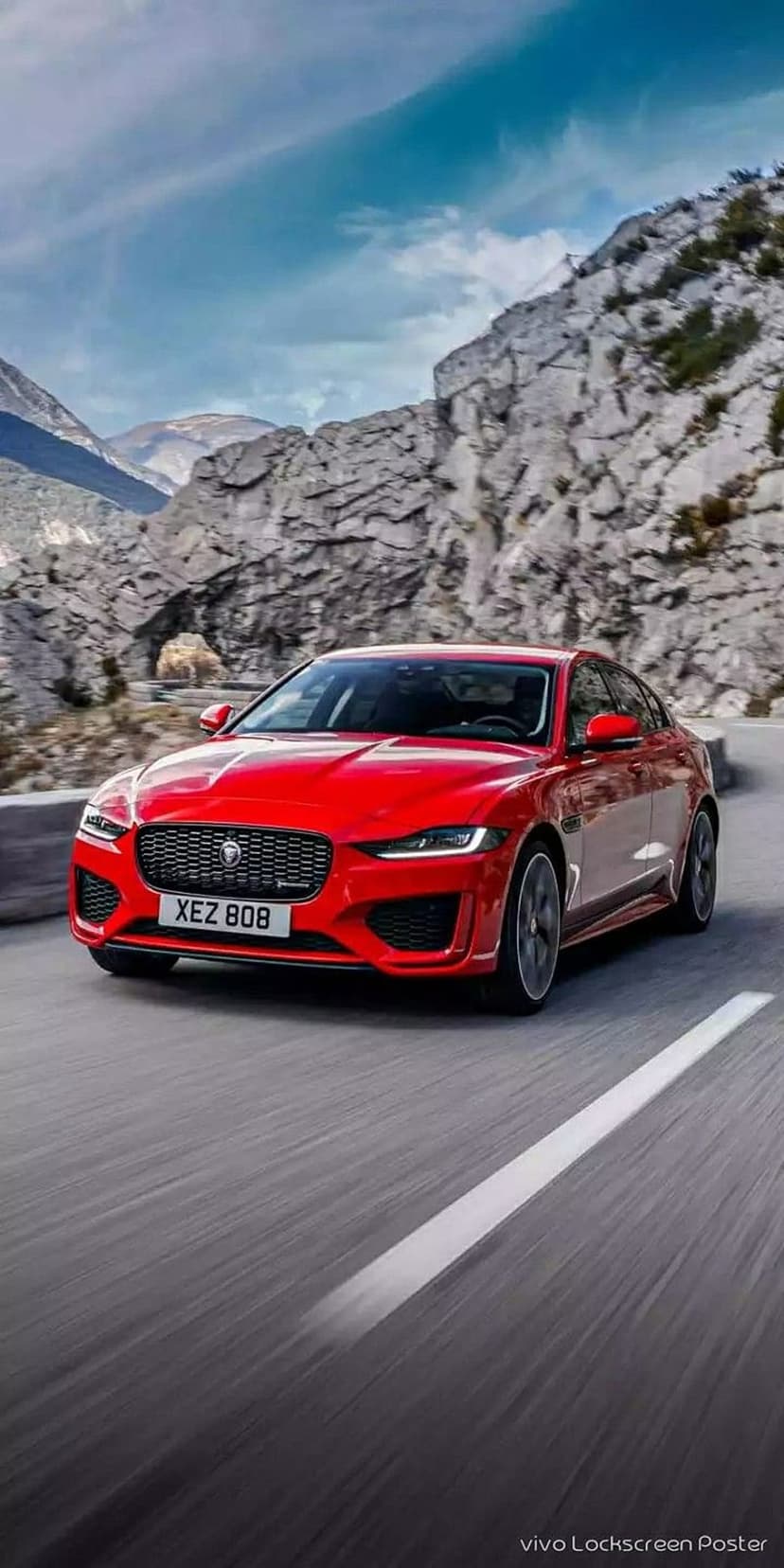 Red Jaguar XE sedan driving on a winding asphalt road through steep, rocky mountain cliffs under a cloudy sky.