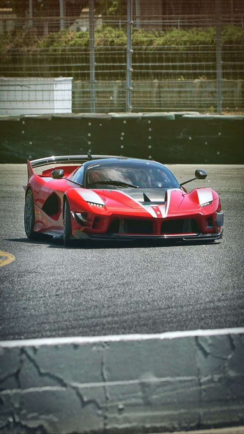 Red Ferrari FXX K Evo supercar on an asphalt race track with a white racing stripe and green blurred background.