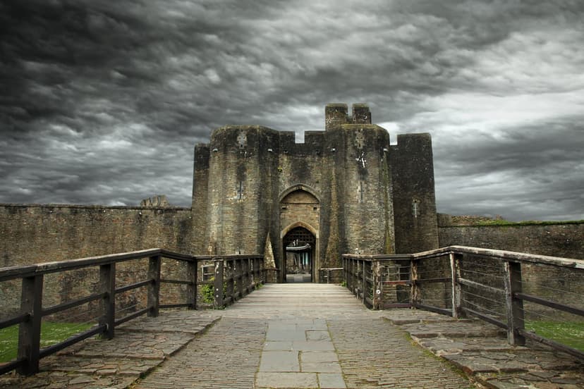 Ancient stone castle gatehouse with a wooden bridge stands under a dark, swirling sky of stormy grey clouds.
