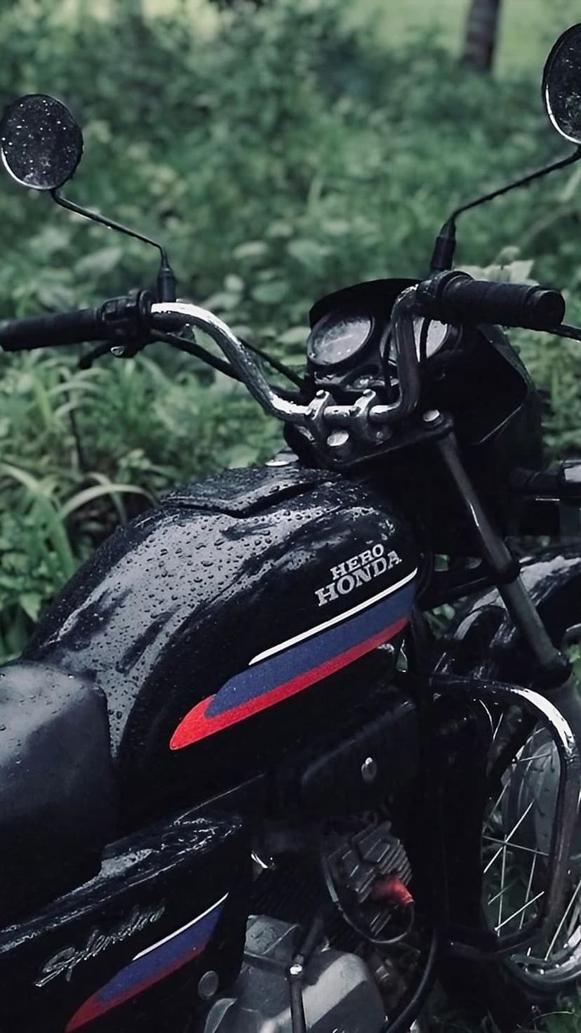 Hero Honda Splendor motorcycle with raindrops on its fuel tank parked against a backdrop of green foliage.