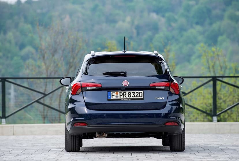 Rear of a dark blue Fiat Tipo wagon parked on a paved road against a blurred green hillside.