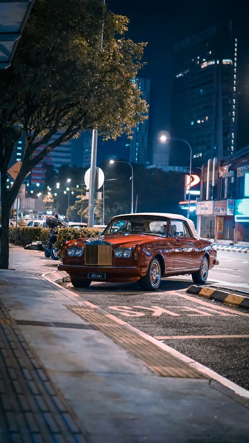 Red vintage Rolls-Royce convertible parked on a wet city street at night with glowing skyscraper windows behind.
