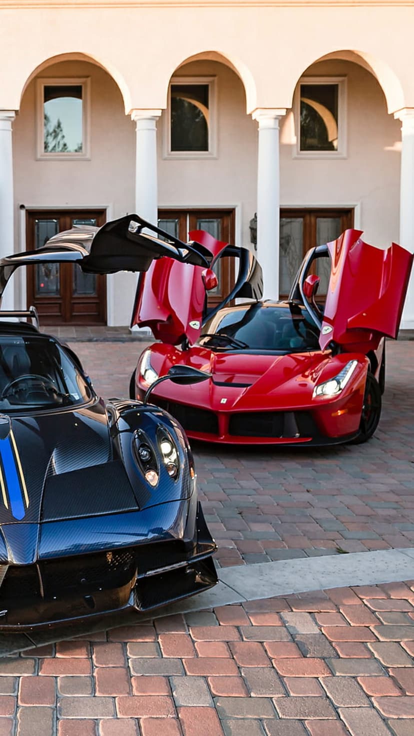 Blue Pagani and red Ferrari hypercars with doors open parked on a stone driveway before a majestic mansion.