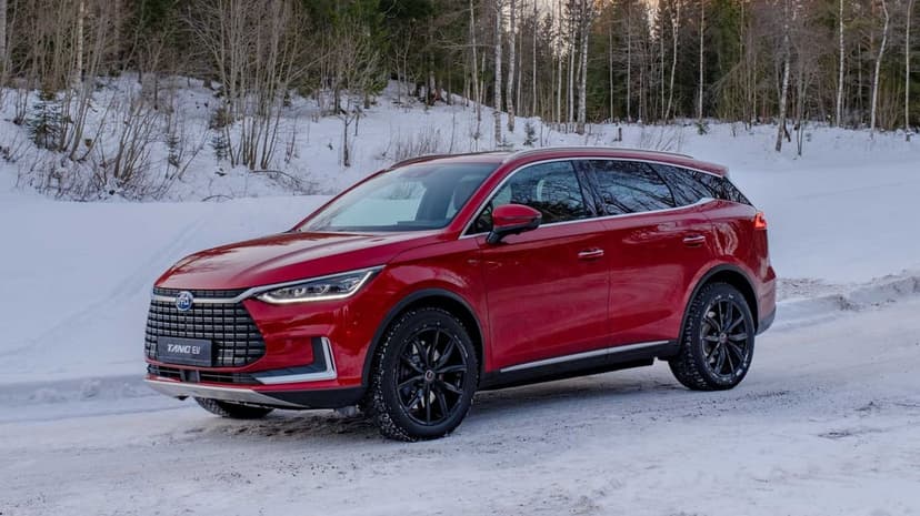 Red BYD Tang EV parked on a snowy road with a dense forest of leafless trees under soft winter light.