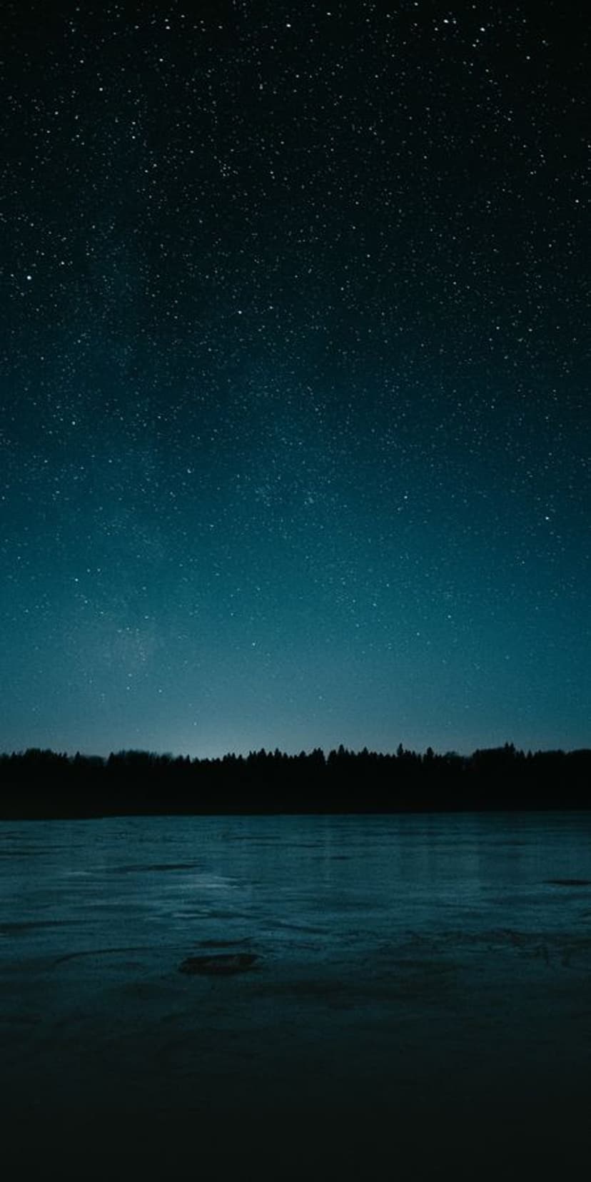 Starry night sky with a shimmering galaxy reflects in a still lake bordered by a dark forest silhouette.