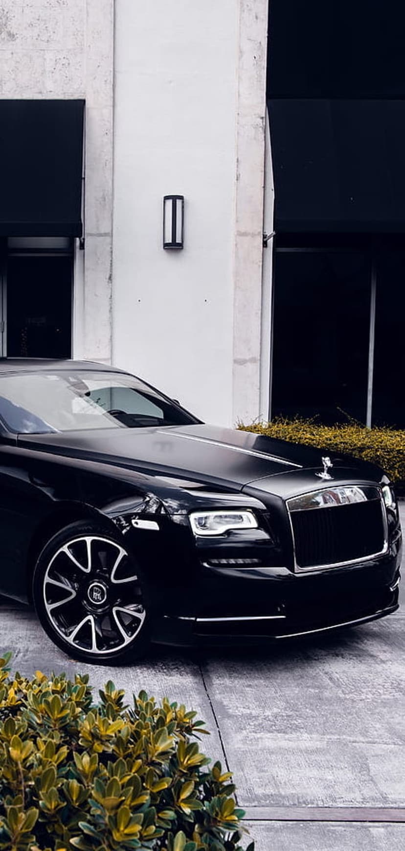 Black Rolls-Royce Wraith luxury car parked near a contemporary glass building with manicured green hedges.