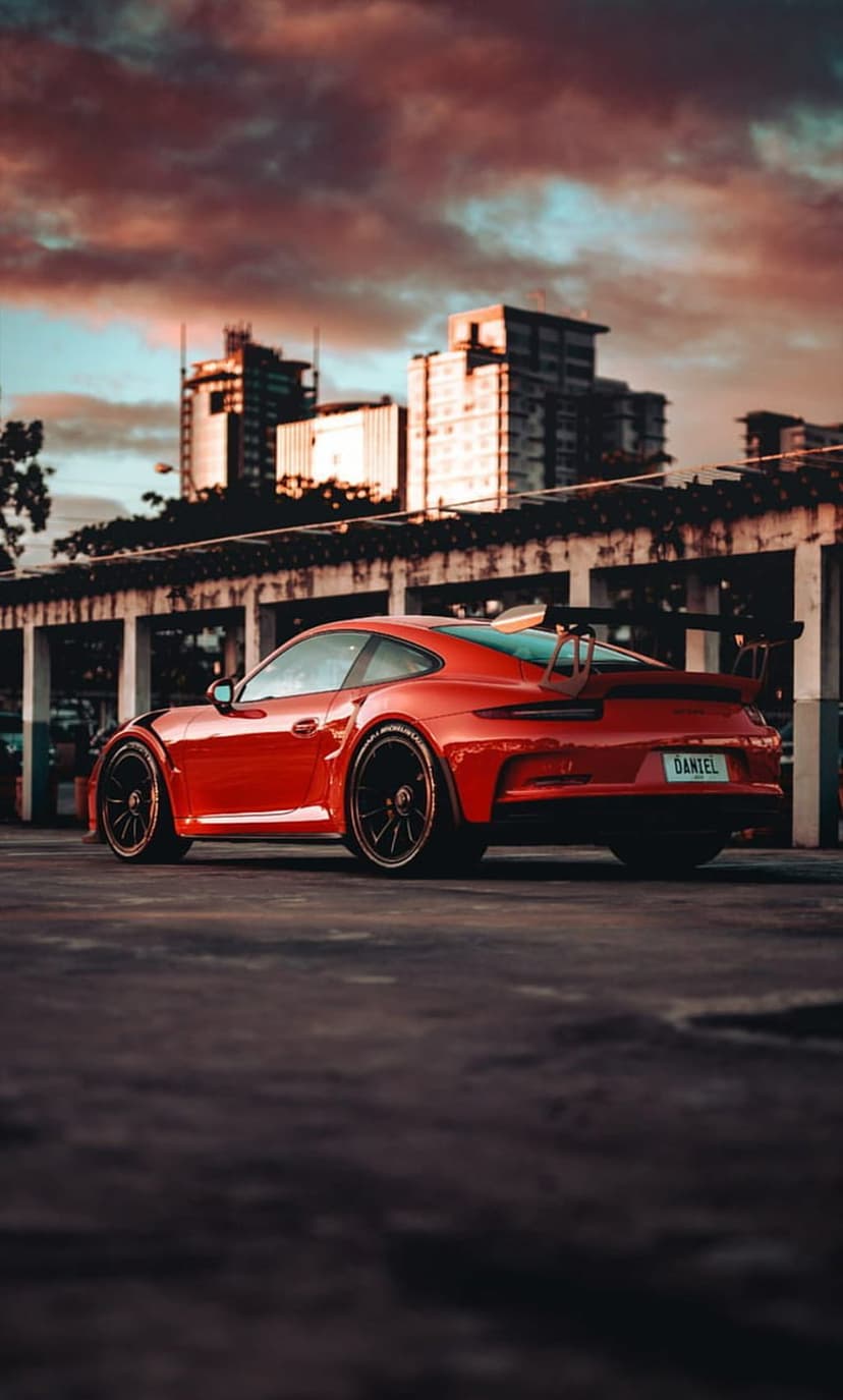 Red Porsche 911 GT3 with a large rear spoiler parked on asphalt during a vivid sunset with a city background.