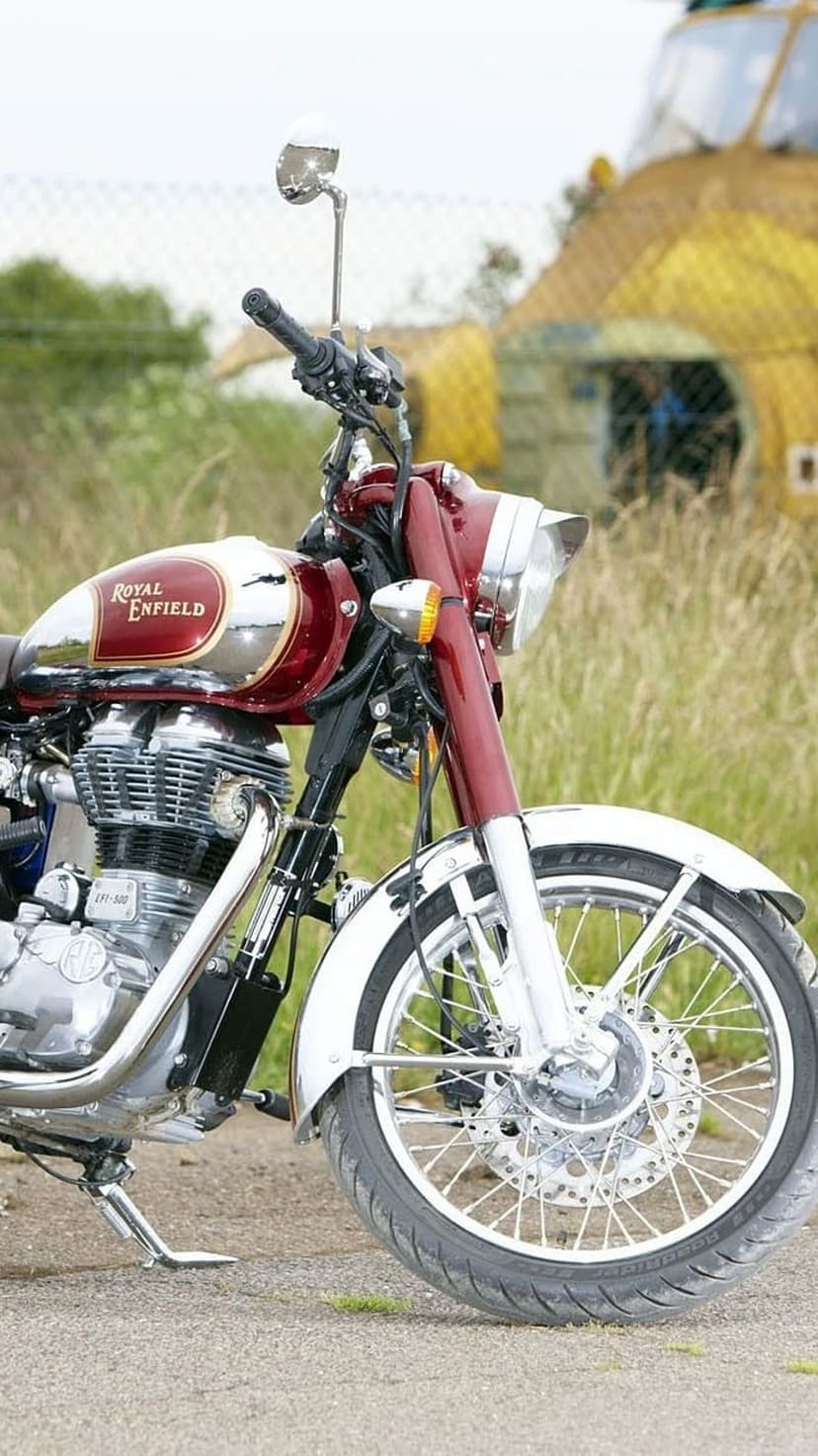 Royal Enfield motorcycle with a maroon fuel tank and chrome engine components parked on a grey gravel surface.