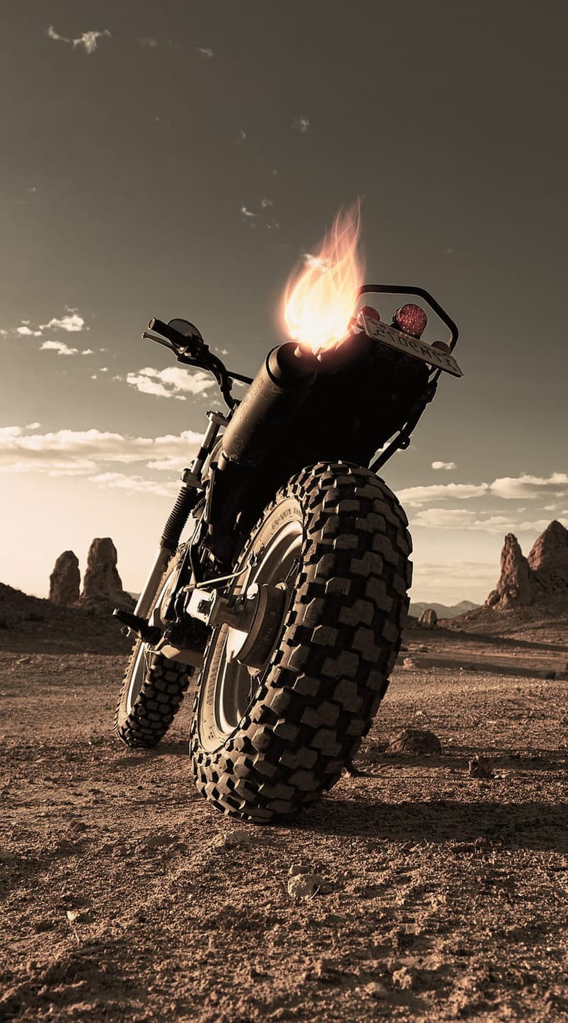 Motorcycle exhaust pipe emitting bright orange flames on a sandy desert trail under a cloudy grey sky background.