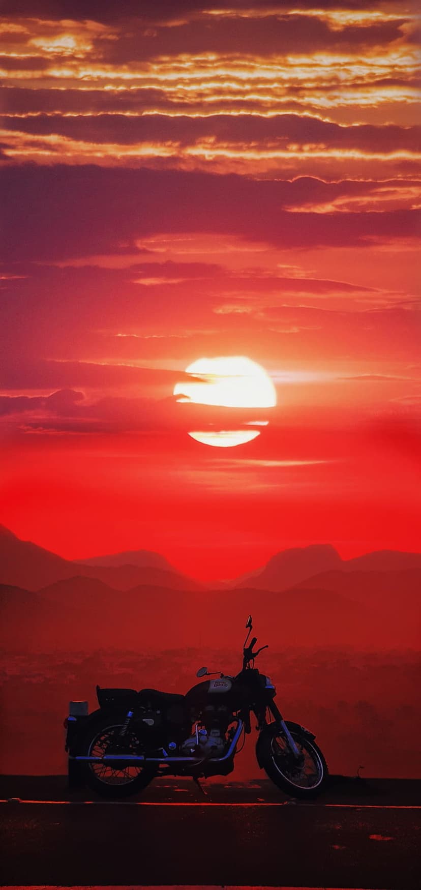 Motorcycle silhouette on a mountain ridge under a fiery red and orange sunset sky with distant layered peaks.